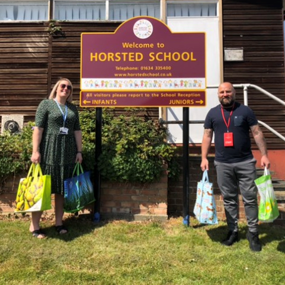 A man and a woman standing by a sign for Horsted School and holding bags of food.