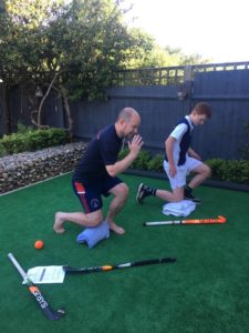 A man and teenage boy in a garden doing fitness exercises with hockey sticks and balls lying on the ground next to them.