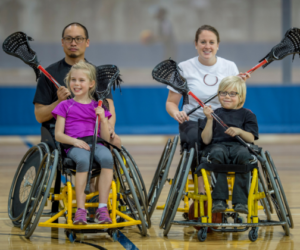 Two adults and two children in wheelchairs in an indoor sports hall. They are holding lacrosse sticks and looking at the camera.