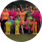 Crowd of people in colourful orange, pink and green t shirts standing on a grass field in front of some cricket stumps and bats.